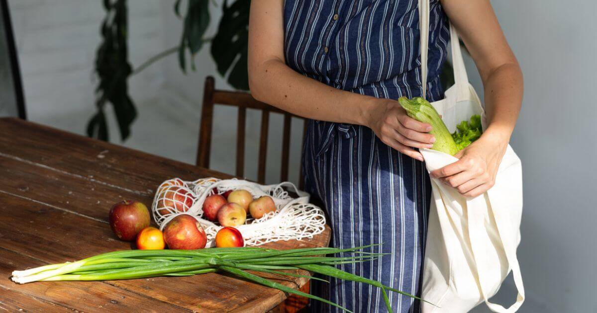 Someone in a blue stripy jumpsuit holing a cloth tote bag and putting vegetables and fruit in it