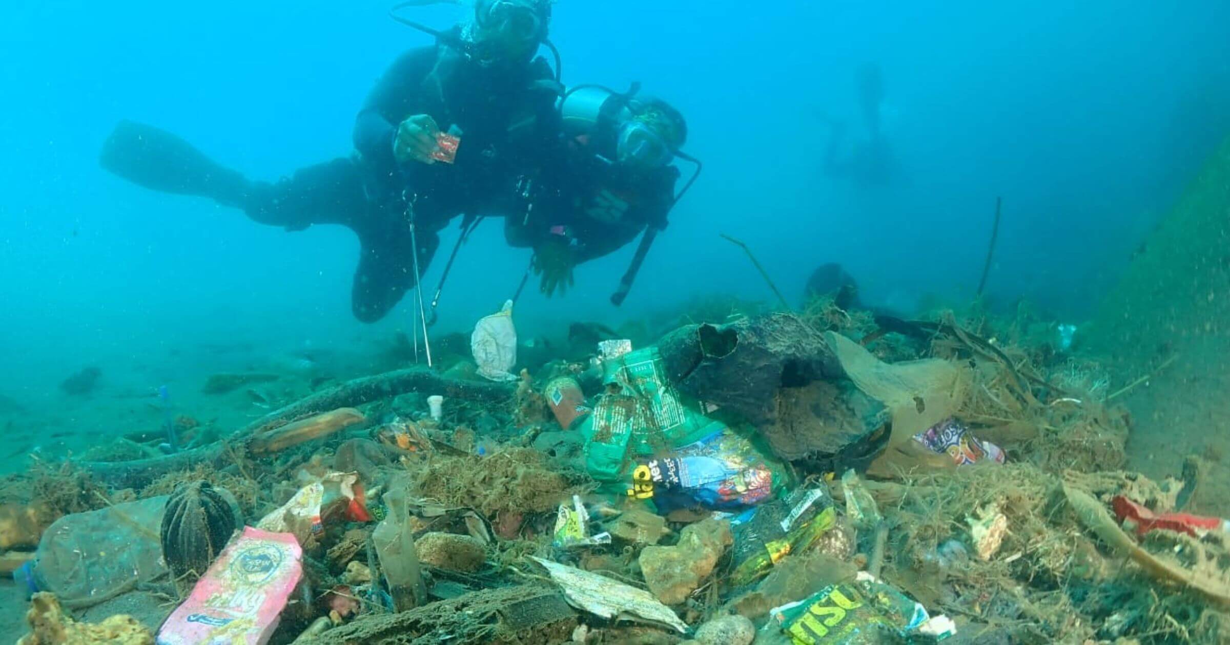 Two divers hover above piles of plastic items on the ocean floor