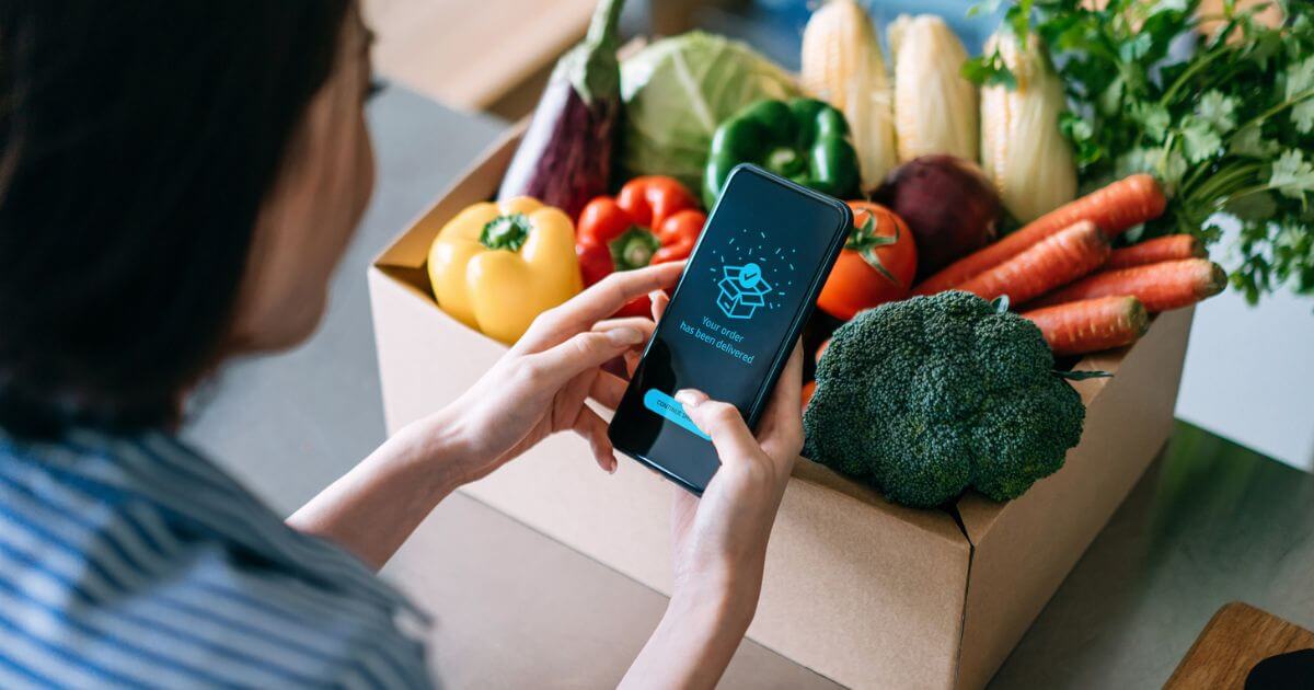 Someone holding their phone over a box of unwrapped vegetables in a cardboard box