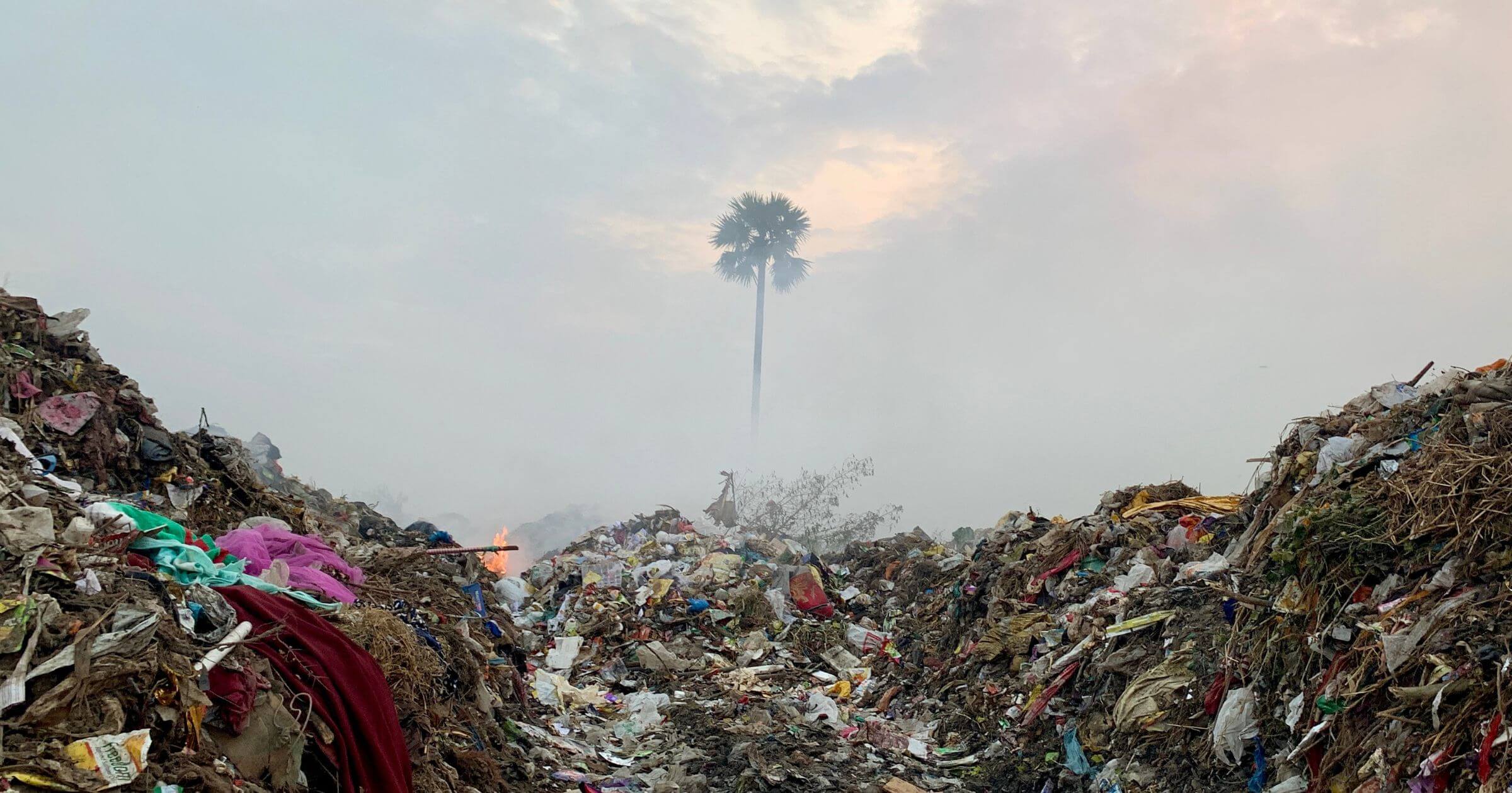 Piles of trash in a landfill with a fire in the distract and a smokey sky