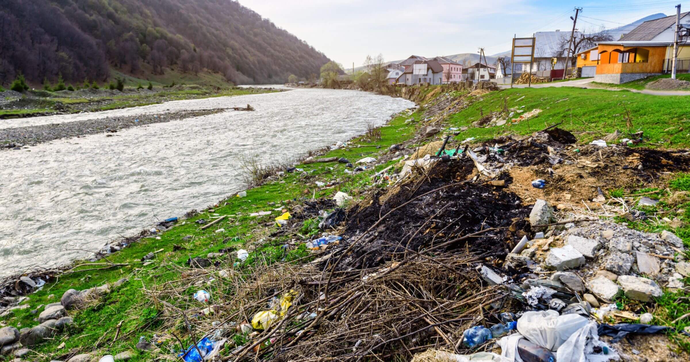Plastic items washed up on a riverbed with houses and a hilltop in the background