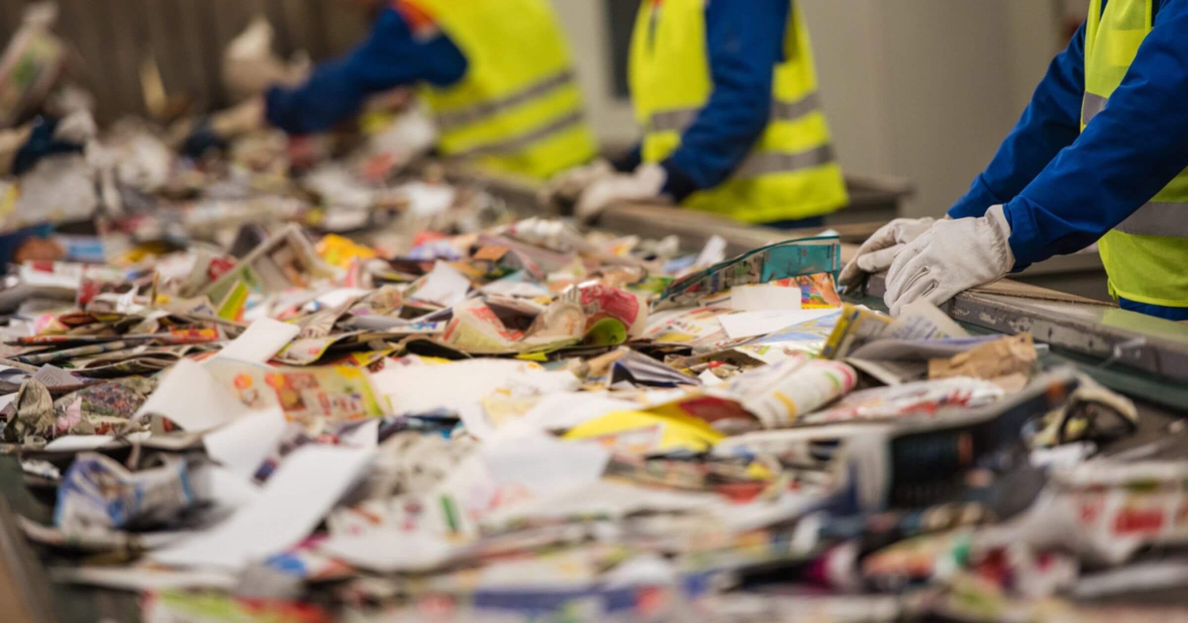 Lots of paper and cardboard laid out on a conveyer belt with people in high viz jackets sorting it
