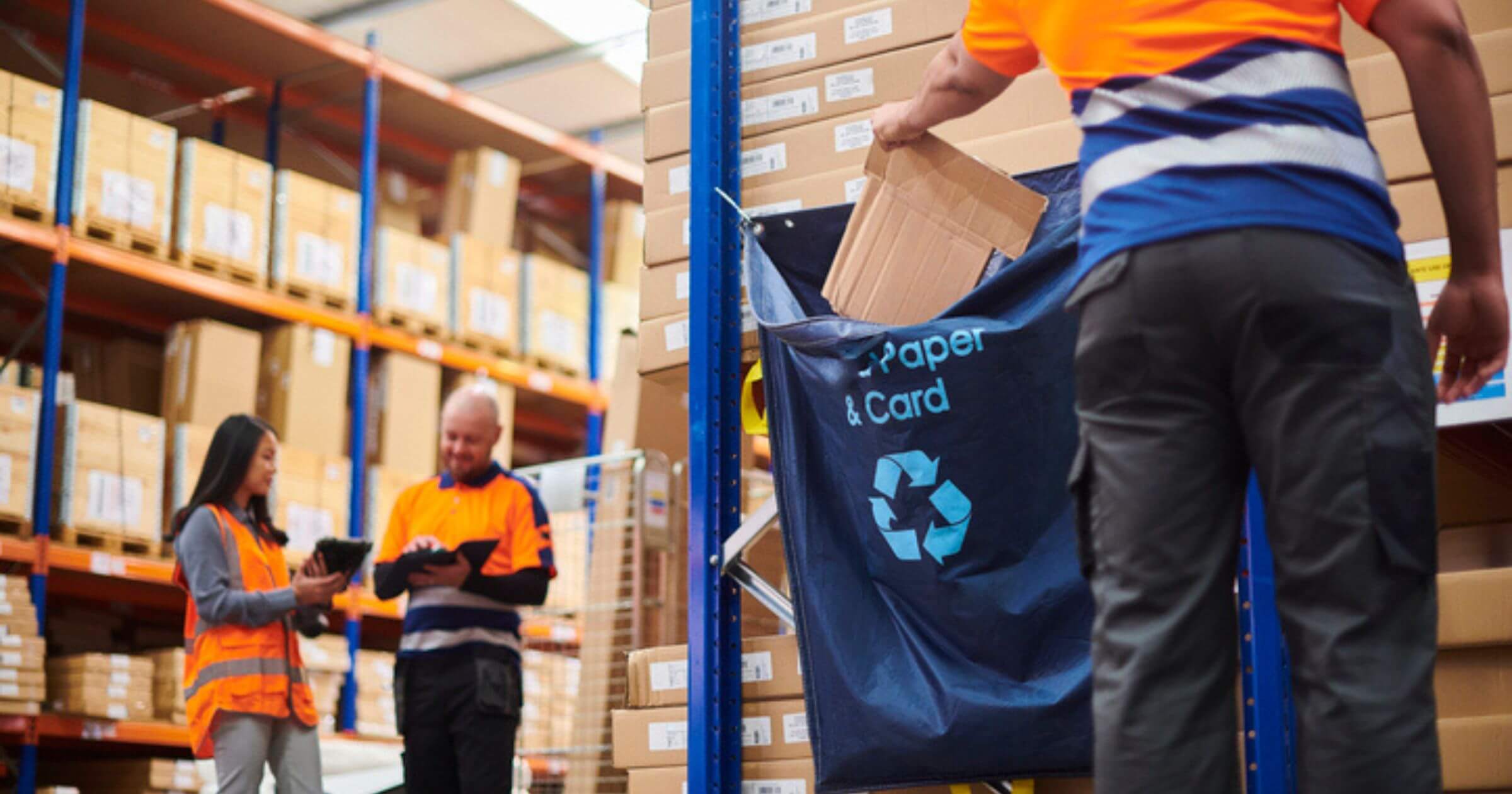 Three people in a warehouse with one person in the foreground putting cardboard in a recycling bag