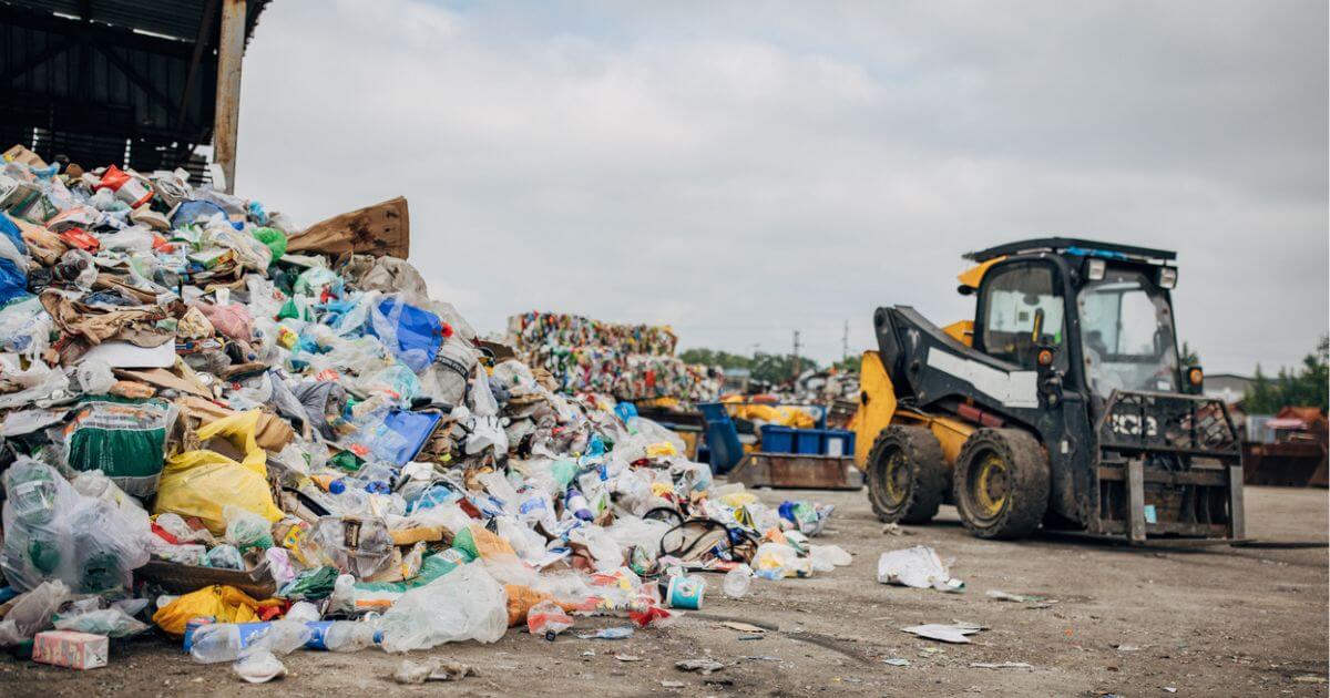 A large pile of trash in a sorting facility with a digger driving past