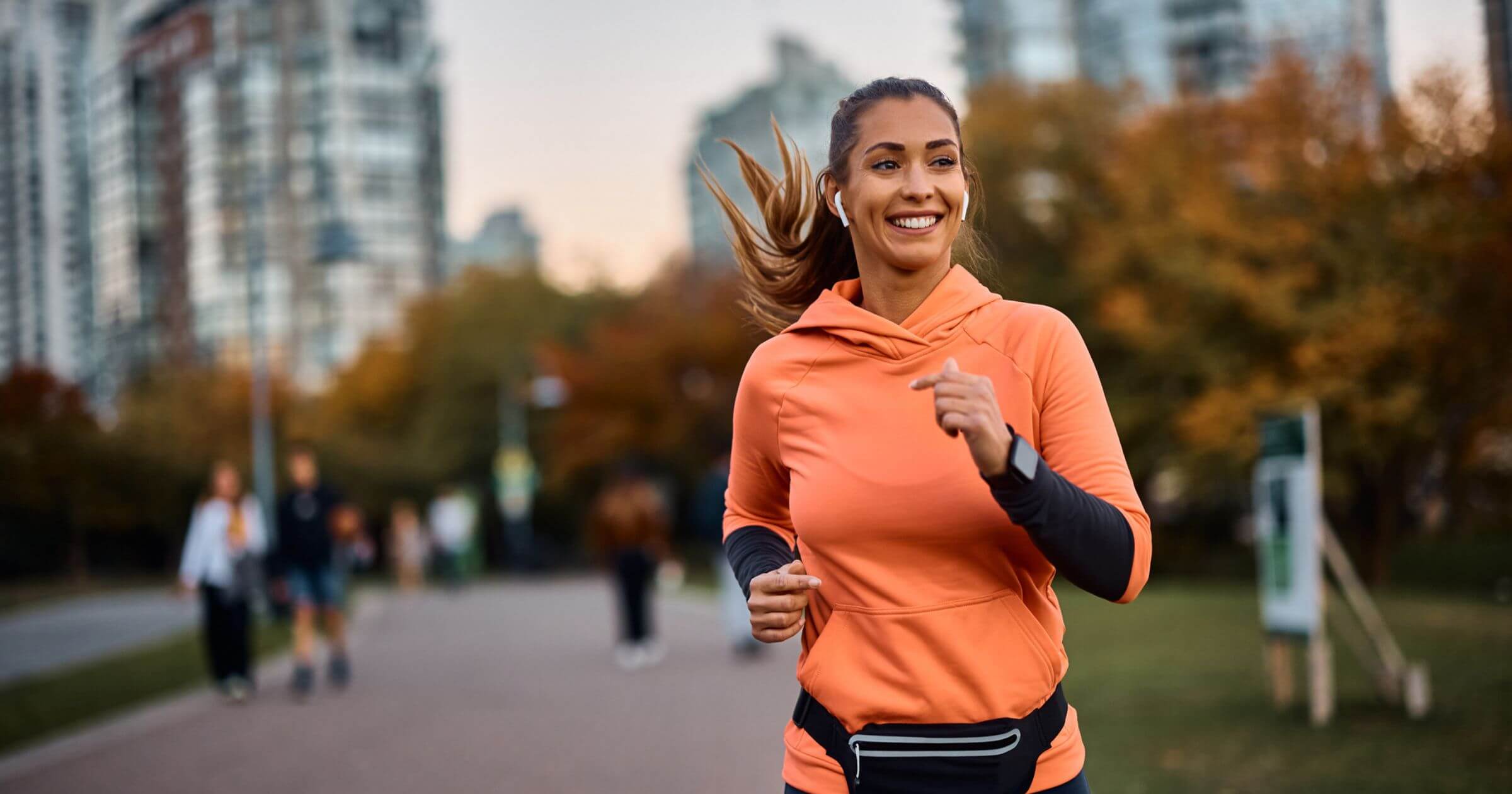 A woman smiling wearing an orange jumper as she runs through a park in a city